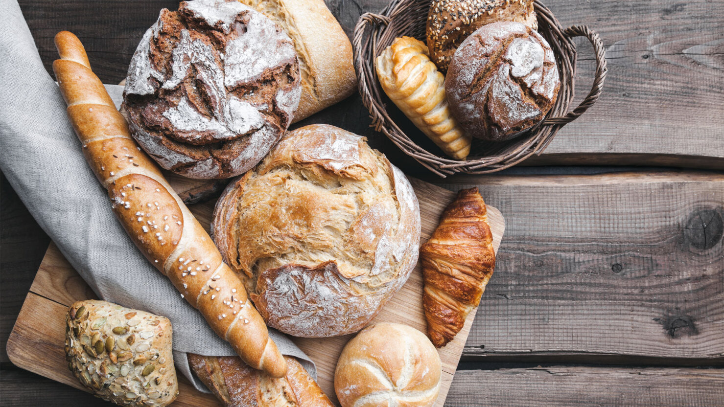 Assorted breads, including baguettes, round loaves, croissants, and rolls, arranged on a wooden surface and in a basket, with a linen cloth partially covering some bread.