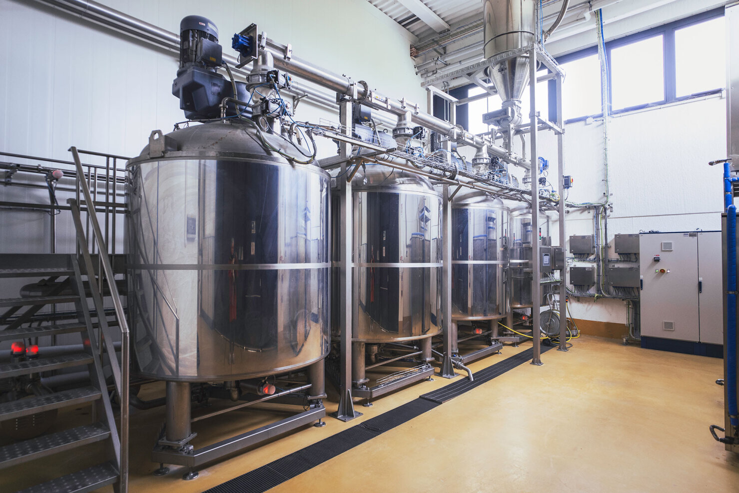 Three large stainless steel industrial tanks with pipes and machinery in a clean, well-lit factory setting. Control panels and metal stairs are visible, suggesting a processing or manufacturing facility.