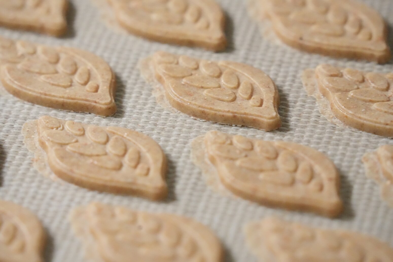 Several leaf-shaped cookies with a patterned design are arranged in rows on a silicone baking mat, ready to be baked.