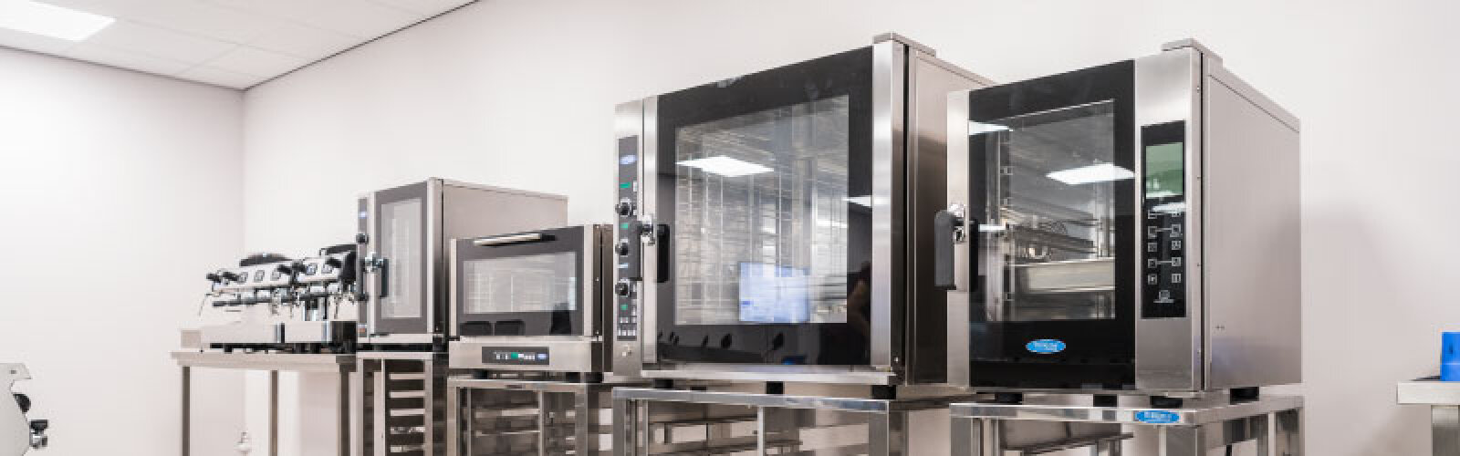 Eine Reihe moderner gewerblicher Küchengeräte aus Edelstahl, darunter Backöfen und eine Espressomaschine, angeordnet auf Metalltischen in einer hellen, sauberen Küche.