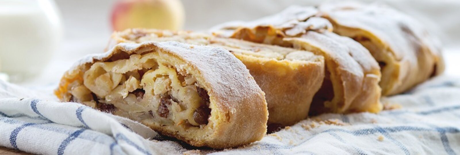 Eine Nahaufnahme eines aufgeschnittenen Apfelstrudels mit einer goldenen, blättrigen Kruste und einer Füllung aus Äpfeln und Rosinen, bestäubt mit Puderzucker, der auf einem gestreiften Tuch liegt.