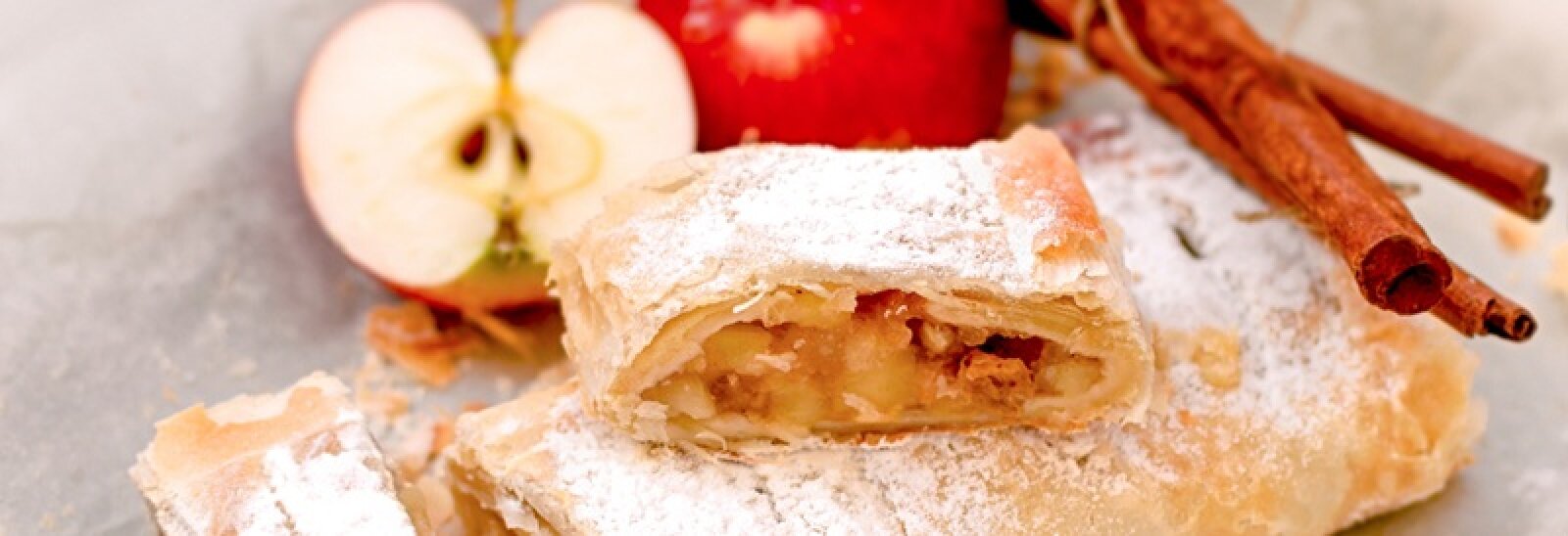 Zu sehen sind zwei mit Puderzucker bestäubte Apfelstrudelstücke. Im Hintergrund liegen ein halbierter roter Apfel und zwei Zimtstangen auf einer hellen Unterlage.