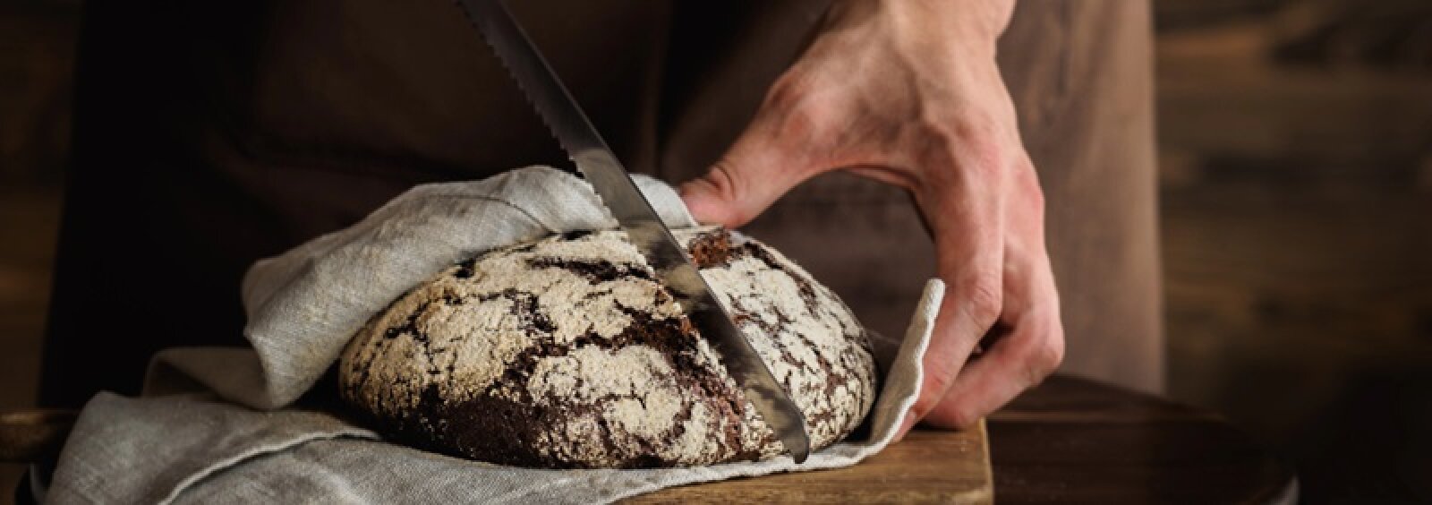 Eine Person schneidet einen runden, rustikalen Laib Brot mit einem gezackten Messer und hält das Brot mit einem Tuch auf einer Holzoberfläche fest.
