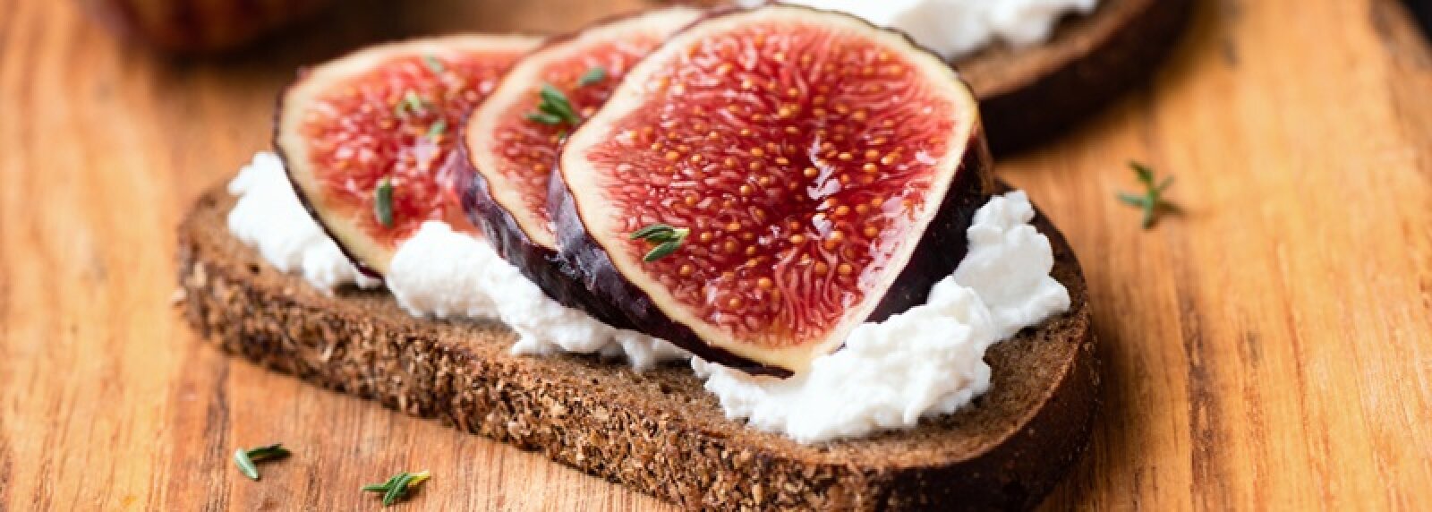 Eine Scheibe dunkles Brot mit weißem Ricotta und drei Scheiben frischer Feige, garniert mit kleinen grünen Kräutern, auf einer Holzoberfläche.