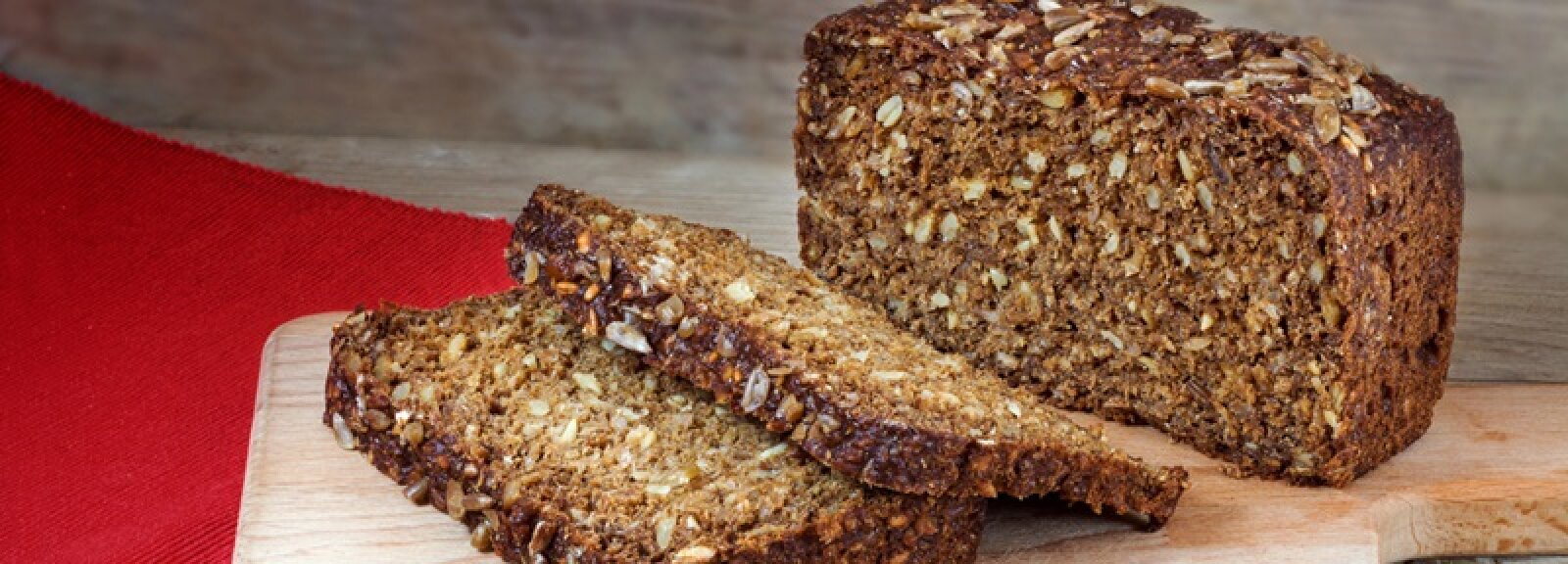 Auf einem hölzernen Schneidebrett liegt ein Laib Vollkornbrot mit Körnern. Vor ihm liegen zwei angeschnittene Scheiben, daneben liegt ein rotes Tuch.