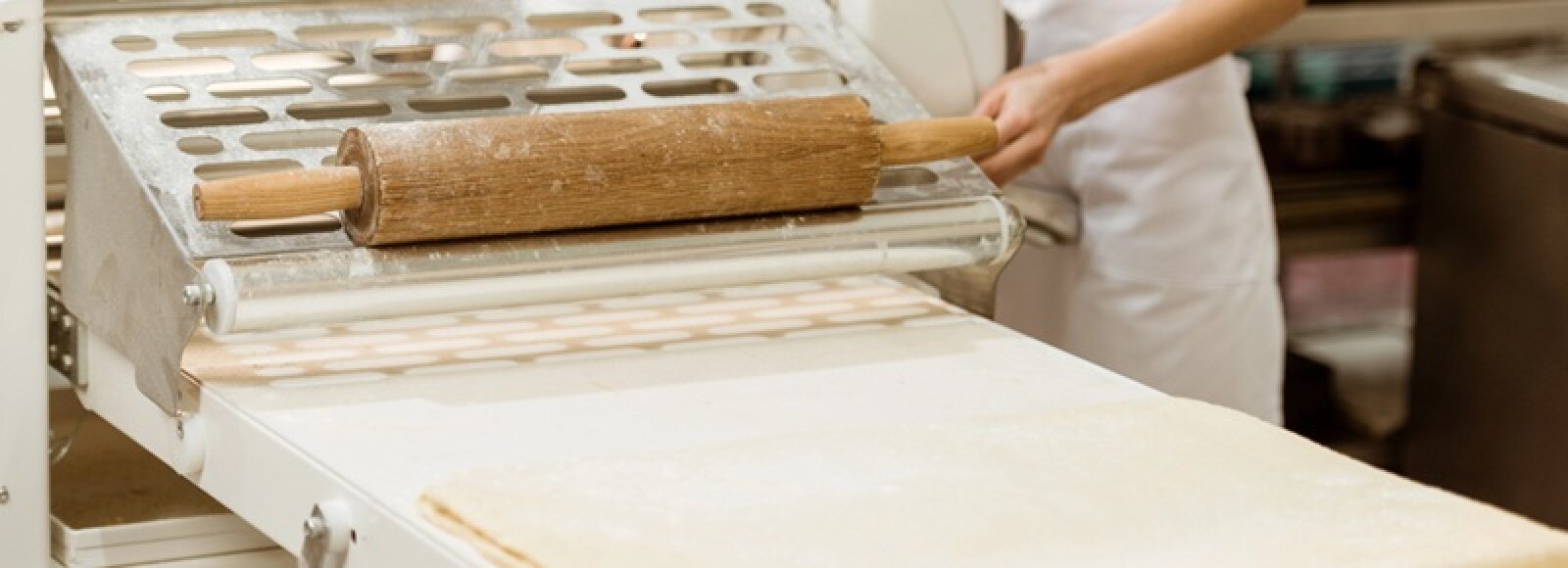 Eine Person in einer weißen Schürze hält ein hölzernes Nudelholz über eine Metalloberfläche und bereitet in einer Großküche oder Bäckerei Teig zu.