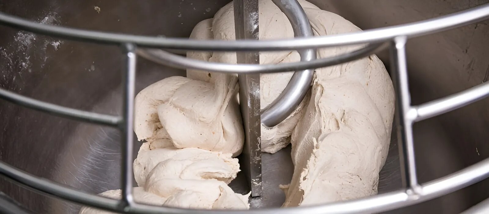 Dicker Brotteig wird in einer industriellen Küchenmaschine aus Metall mit einem Spiralhaken geknetet. Im Vordergrund ist teilweise ein Schutzgitter aus Metall zu sehen.